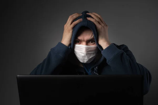 Den maskerade mannen bakom laptopen och i chock läser nyheter om världshändelser och pandemin. — Stockfoto