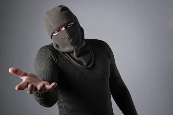 Um homem em uma balaclava em um fundo cinza está, segurando a mão — Fotografia de Stock