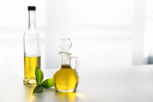 Tegen het raam, olijfolie in een karaf en in een glazen fles, met een groen blad — Stockfoto