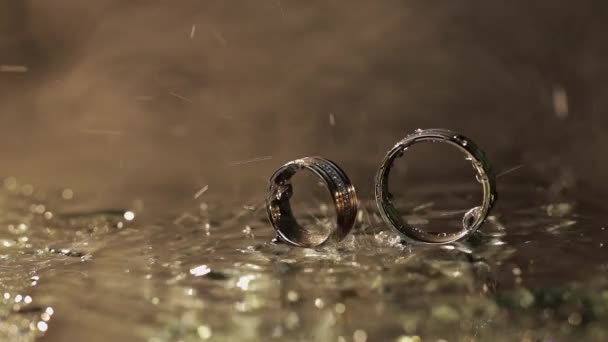 Свадебные кольца на темной поверхности воды сияют светом. Капли воды. Закрыть — стоковое видео