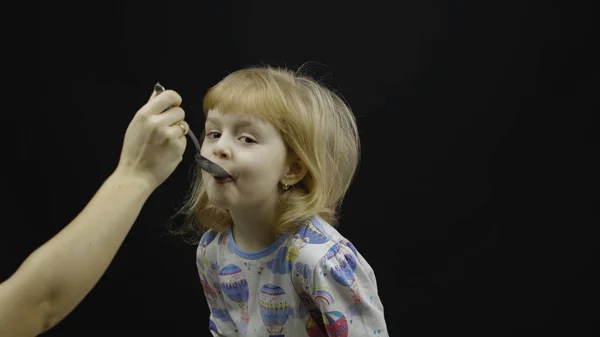 Een klein meisje in pyjama eet pap uit de lepel. Zwarte achtergrond — Stockfoto