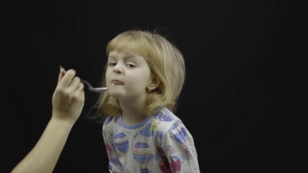 En liten flicka i pyjamas äter gröt från skeden. Svart bakgrund — Stockvideo