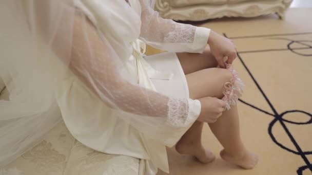Menyasszony lábai hálóingben és fátyolban. Ülni egy kanapén és harisnyakötőt tenni a lábára. — Stock videók