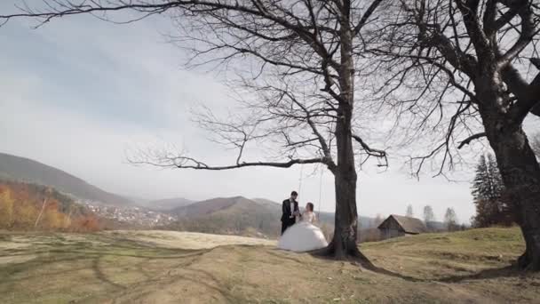 新婚旅行だよ。山の斜面にロープスイングに乗る花嫁と白人新郎 — ストック動画