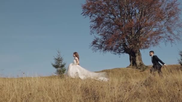 Recién casados. novio caucásico con novia corriendo en la ladera de la montaña. Pareja de boda — Vídeo de stock