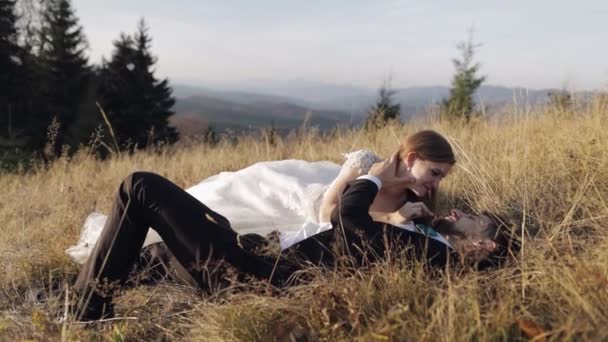 Newlyweds. Caucasian groom with bride lie on the grass on mountain slope — Stock Video