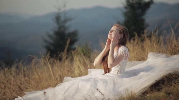 산비탈에 있는 풀 위에 앉아 있는 아름답고 아름다운 웨딩 드레스를 입은 신부 — 비디오