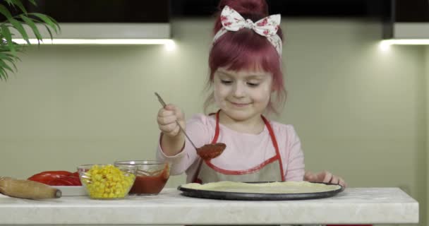 Cocinar pizza. Niño pequeño en delantal añadiendo salsa de tomate a la masa en la cocina — Vídeo de stock