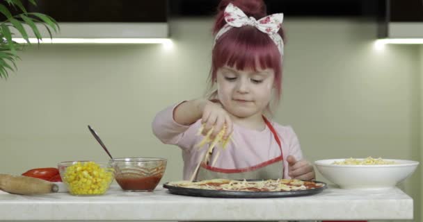 Vařím pizzu. Malé dítě v zástěře přidání strouhaného sýra do těsta v kuchyni — Stock video