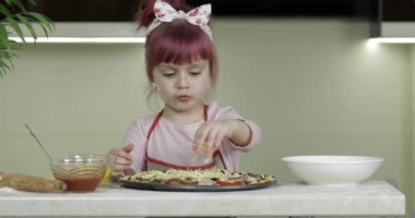 Yemek pişirmek. Önlüklü küçük çocuk mutfakta çiğ pizzaya rendelenmiş peynir ekliyor.