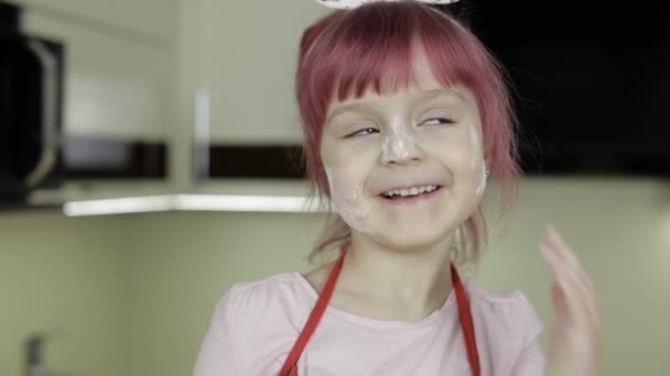 Маленька дівчинка на кухні, одягнена в фартух і шарф. Обличчя в борошні — стокове відео