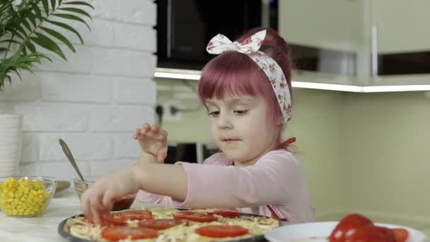 Cooking pizza. Little child in apron adding sliced tomatoes to dough in kitchen — Stock Video