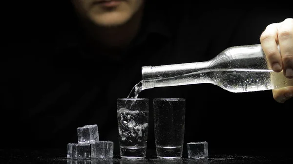 Il barman versa la vodka congelata da bottiglia in due occhiali con ghiaccio. Fondo nero Fotografia Stock