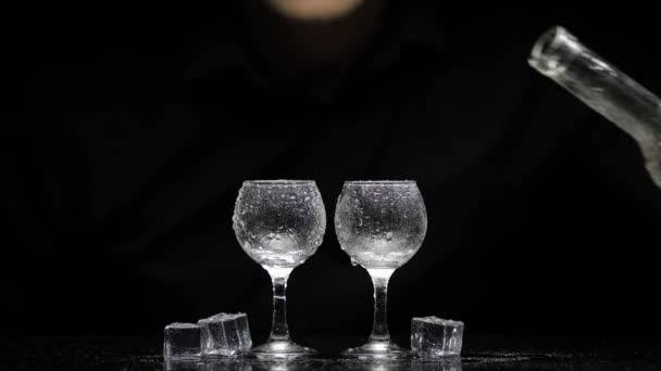 Barmann gießt gefrorenen Wodka aus der Flasche in zwei Gläser mit Eis. Schwarzer Hintergrund — Stockvideo