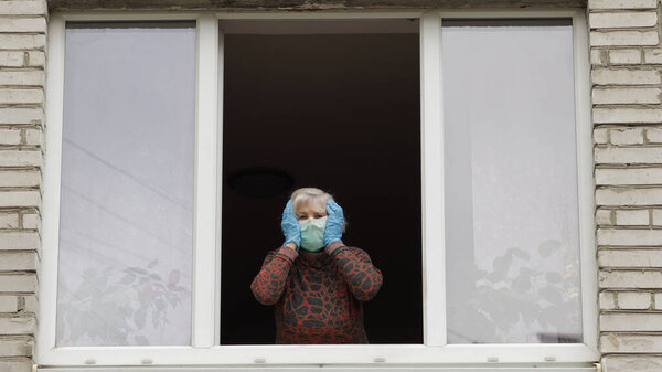 Старуха-бабушка сидит дома в изоляции у окна на карантине. Коронавирус

