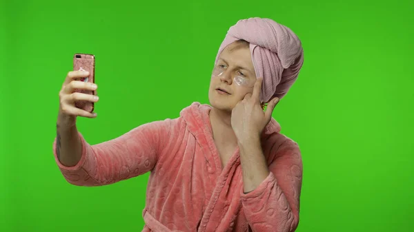 Transsexual man in bathrobe with face mask making selfies using mobile phone — Stock Photo, Image