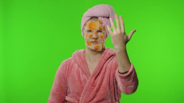 Transexual hombre en albornoz con máscara facial ondea su mano, dando pulgares hacia arriba — Vídeos de Stock