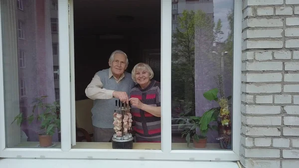 Old grandparents couple cooking barbecue on electric grill on windowsill at home