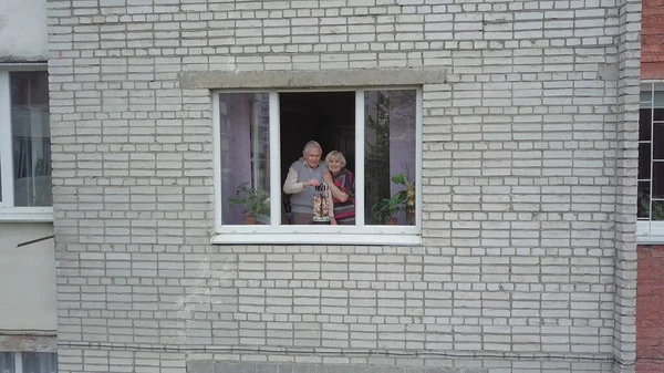 Old grandparents couple cooking barbecue on electric grill on windowsill at home