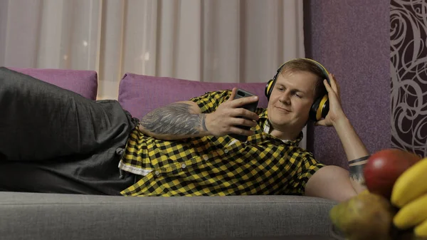 ソファの上に横たわる男は、ワイヤレスヘッドフォンでスマートフォンから音楽を聞く — ストック写真