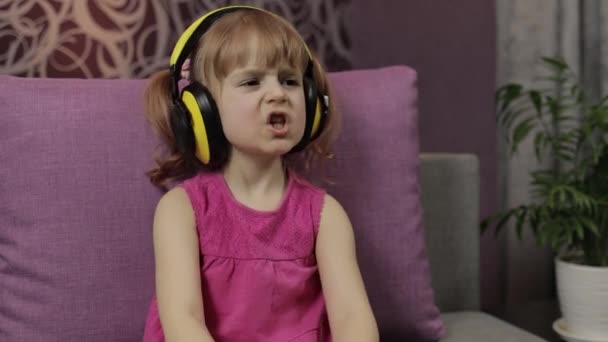ヘッドフォンで音楽を聞いて楽しむ小さな子供の女の子。自宅でソファで踊る — ストック動画