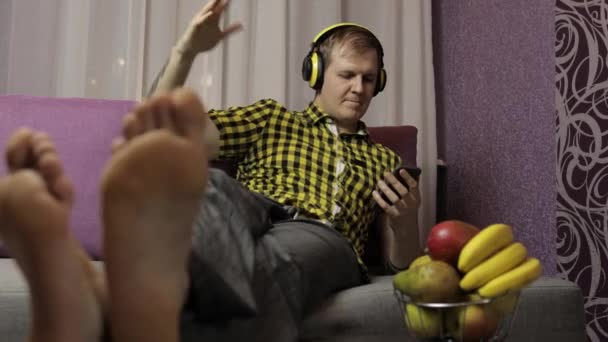 Człowiek leżący na kanapie słucha muzyki bezprzewodowymi słuchawkami i tańcami — Wideo stockowe