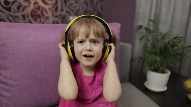 Menina pequena em fones de ouvido apreciando ouvir música. Dançando no sofá em casa — Vídeo de Stock