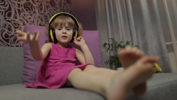 Menina pequena em fones de ouvido apreciando ouvir música. Dançando no sofá em casa — Vídeo de Stock