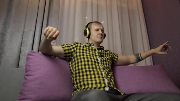 Мужчина, сидящий на диване, слушает музыку из беспроводных наушников и танцев — стоковое видео