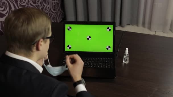 Mand fjerner medicinsk maske, tager desinfektionsmiddel og bruger nær bærbar computer med grøn skærm – Stock-video
