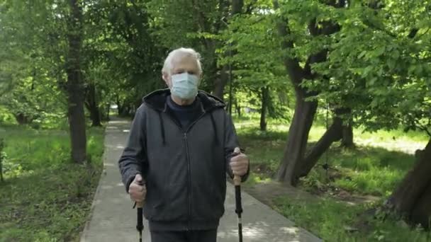 Активный пожилой человек в маске тренирует нордическую ходьбу в парке во время карантина — стоковое видео