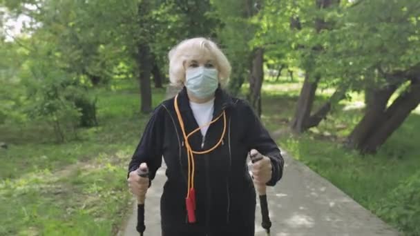 Активна старша жінка в масці тренує скандинавську ходьбу в парку в карантині — стокове відео