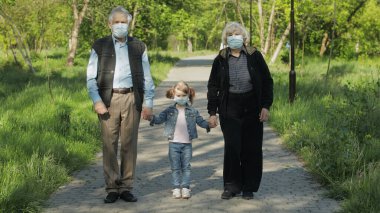 Tıbbi maskeli torunları olan yaşlı büyükanne ve büyükbabaları parkta yürürler. Coronavirüs