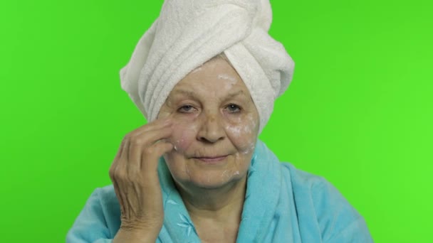 Bornozlu yaşlı büyükanne. Yaşlı kadın yüzüne nemlendirici krem sürüyor. — Stok video