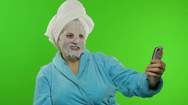 Nagymama fürdőköpenyben, arcmaszkban. Öreg nő csinál szelfik segítségével mobiltelefon — Stock Fotó