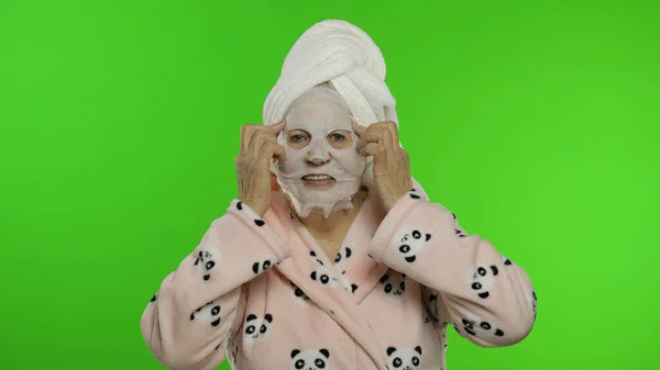 Abuela mayor después de la ducha. Mujer vieja aplicando máscara facial de tela cosmética —  Fotos de Stock