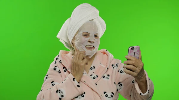 Grandmother in bathrobe, face mask. Old woman making selfies using mobile phone — Stock Photo, Image