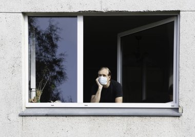 Tıbbi koruyucu maskeli bir adam pencereden bakıyor. Coronavirus pandemik covid-19.Adam karantinada pencereden sokağa bakıyor. Yüz maskeli adam evde kilit altında.