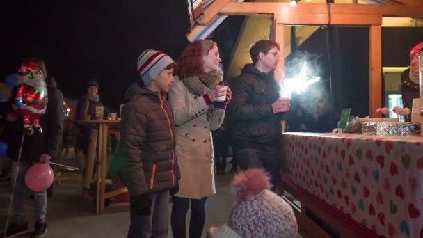 两个成年人站在一个圣诞摊前 手里拿着含酒精的葡萄酒 他们的孩子和他们在一起 — 图库视频影像