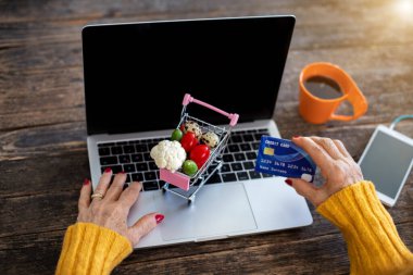 Yaşlı bir kadın kredi kartıyla online alışveriş yapıyor. Özellikle Coronavirus 'tan sonra alışveriş arabasıyla ticaret konsepti.