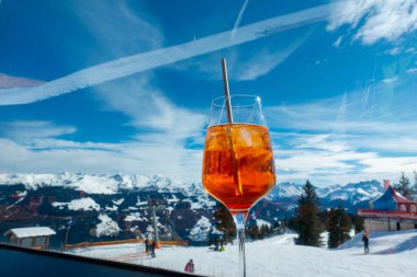 Bir masada portakallı uzun bir içecek bardağı ve arka planda karla kaplı dağlar var. 