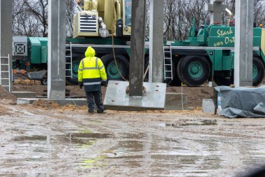 Sarı yağmurluklu bir inşaat işçisi büyük bir beton sütunun ereksiyonunu denetliyor ve kontrol ediyor.