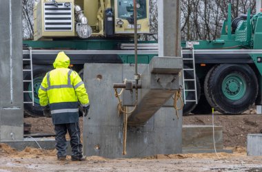 Sarı yağmurluklu bir inşaat işçisi büyük bir beton sütunun ereksiyonunu denetliyor ve kontrol ediyor.