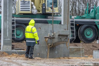 Sarı yağmurluklu bir inşaat işçisi büyük bir beton sütunun ereksiyonunu denetliyor ve kontrol ediyor.