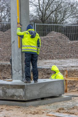 Sarı yağmurluklu bir inşaat işçisi büyük bir beton sütunun ereksiyonunu denetliyor ve kontrol ediyor.