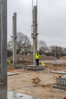 Sarı yağmurluklu bir inşaat işçisi büyük bir beton sütunun ereksiyonunu denetliyor ve kontrol ediyor.