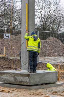 Sarı yağmurluklu bir inşaat işçisi büyük bir beton sütunun ereksiyonunu denetliyor ve kontrol ediyor.