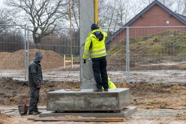 Sarı yağmurluklu bir inşaat işçisi büyük bir beton sütunun ereksiyonunu denetliyor ve kontrol ediyor.
