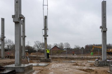 Sarı yağmurluklu bir inşaat işçisi büyük bir beton sütunun ereksiyonunu denetliyor ve kontrol ediyor.