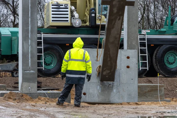 Sarı yağmurluklu bir inşaat işçisi büyük bir beton sütunun ereksiyonunu denetliyor ve kontrol ediyor.